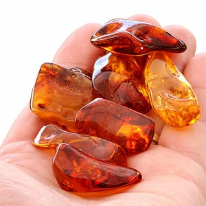 Puede incluir: Una colección de piedras de ámbar pulidas en varios tonos de naranja intenso, amarillo dorado y marrón rojizo. Estas piezas de resina orgánica translúcida presentan bordes suaves y redondeados con formas irregulares.