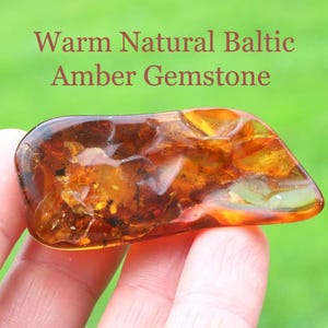 Può includere: Un pezzo levigato e irregolare di ambra del Baltico con tonalità arancioni profonde e dorate, caratterizzato da inclusioni e texture organiche interne. Il testo "Warm Natural Baltic Amber Gemstone" è stampato sopra la pietra.