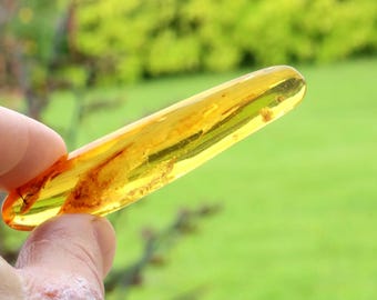 May include: A polished, elongated piece of translucent golden-yellow amber with visible internal organic inclusions and natural texture. The object has a smooth, rounded, tapered shape that catches the light, revealing deep orange and yellow hues.
