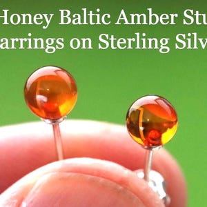 Puede incluir: Pendientes de botón de ámbar báltico color miel sobre plata de ley. Los pendientes presentan piedras redondas de ámbar color miel. El texto "Honey Baltic Amber Stud Earrings on Sterling Silver" se muestra en blanco.