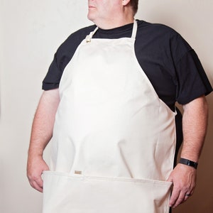 May include: A white canvas apron with a large pocket on the bottom. The apron has a brown strap and a brown buckle. The apron is being worn by a person.