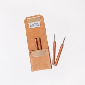 May include: A brown leather case with a beige flap and a label that says "kitchen cozy style". The case contains three wooden tools with metal tips.