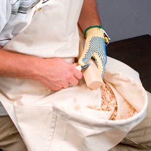 May include: A person wearing a white apron and a blue and yellow dotted glove is using a carving knife to shape a piece of wood. The wood shavings are falling into a white canvas bag attached to the apron.