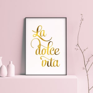May include: A framed print with the text "La dolce vita" in gold script lettering on a white background. The print is displayed on a white shelf with two white vases.