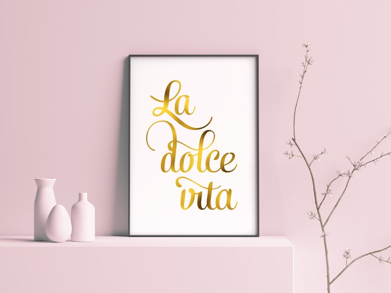 May include: A framed print with the text "La dolce vita" in gold script lettering on a white background. The print is displayed on a white shelf with two white vases.