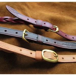 May include: Close-up of three leather straps with brass buckles. The straps are in shades of brown and burgundy, with decorative embossed patterns. One strap is braided. The buckles are gold-coloured.