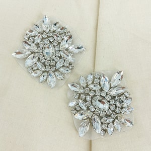May include: Two silver rhinestone embellishments with clear crystals on a white fabric background.