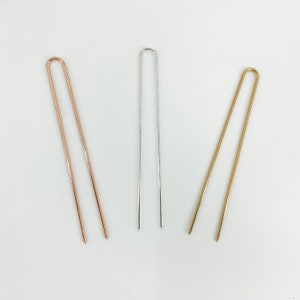 May include: Three metal hair pins in rose gold, silver, and gold. Each pin has a U-shaped top and two straight prongs. The pins are arranged on a white surface.