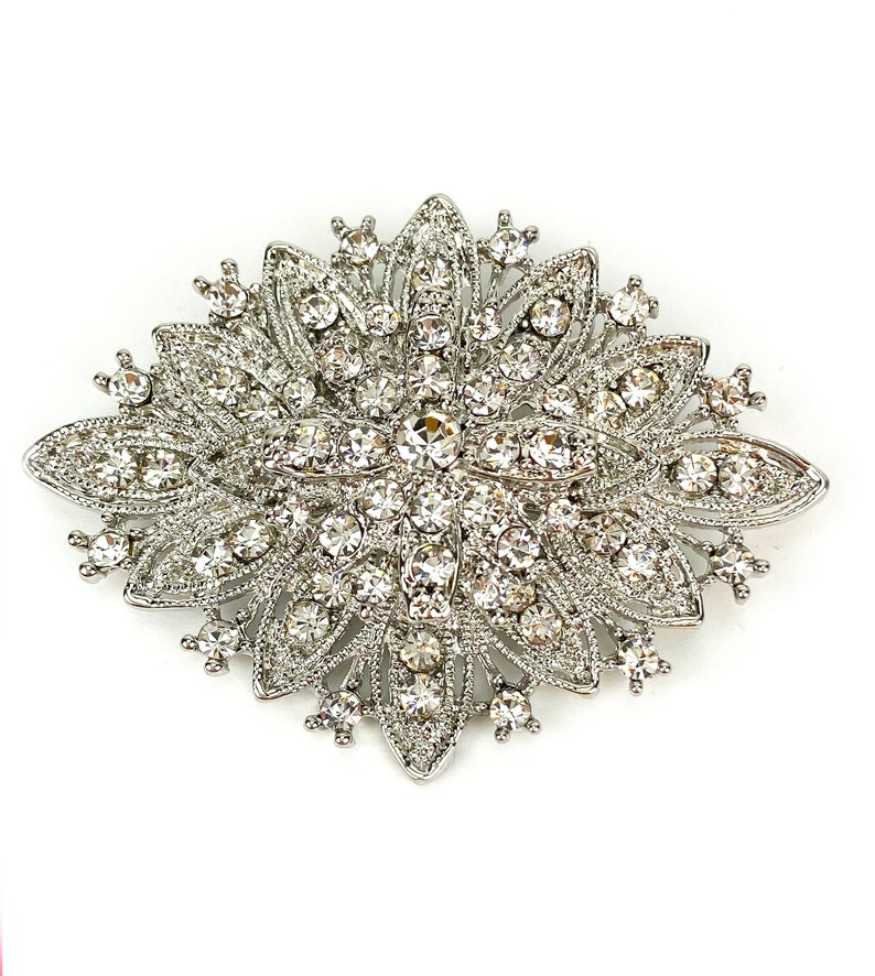 May include: A silver brooch with a large, ornate design featuring a central cluster of clear rhinestones surrounded by smaller rhinestones.