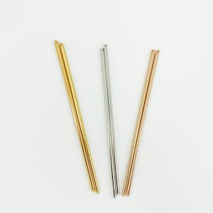May include: Three metal hair sticks in gold, silver, and rose gold. Each stick has a rounded tip and a smooth, polished finish. The sticks are arranged diagonally against a white background.