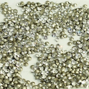 May include: A large quantity of silver-toned, faceted rhinestones with a clear crystal centre. The rhinestones are scattered on a white surface.