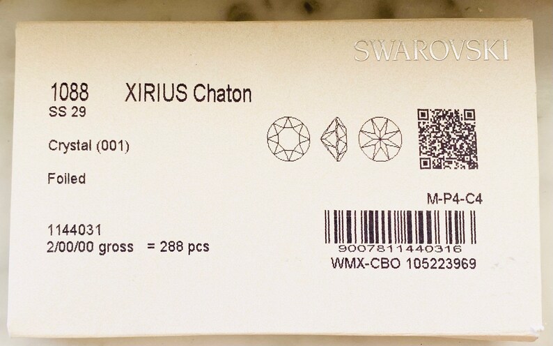 Op de afbeelding: Een close-up van een wit pakket met zwarte tekst die "1088 XIRIUS Chaton SS 29 Crystal (001) Foiled 1144031 2/00/00 gross = 288 pcs" luidt en een barcode. Het pakket bevat ook een kleine zwart-wit afbeelding van een kristal en een QR-code.