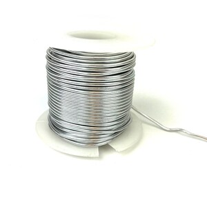 May include: A spool of silver-colored wire on a white plastic spool. The wire is wound tightly around the spool and is ready to be used for crafting or other projects.