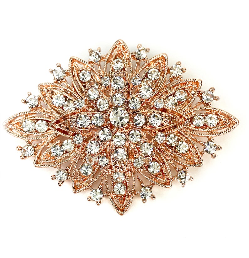May include: A large, rose gold-toned brooch with a floral design. The brooch is covered in clear rhinestones and has a starburst shape.