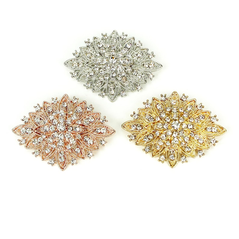 May include: Three decorative brooches with a floral design. The brooches are silver, gold, and rose gold in color and are adorned with clear rhinestones.