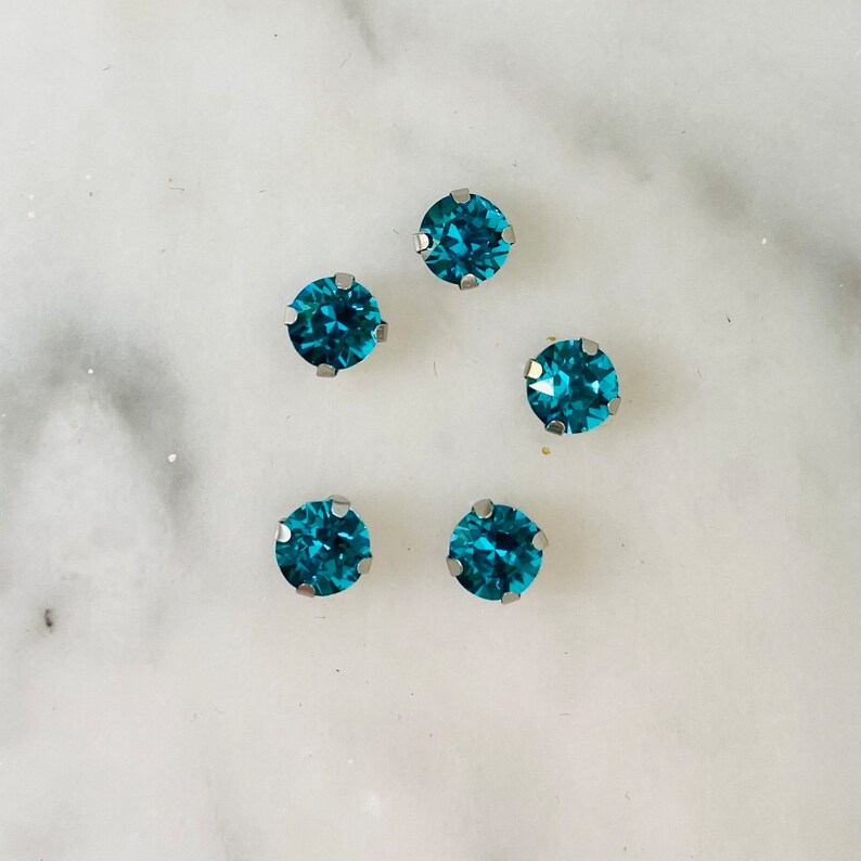 Op de afbeelding: Vijf turquoise blauwe strasssteentjes met zilveren settings. De strasssteentjes zijn in een halve cirkel op een witte achtergrond gerangschikt.