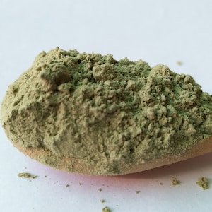 Puede incluir: Una cuchara de madera llena de una buena cantidad de polvo verde. El polvo está finamente molido y tiene una textura ligeramente rugosa.