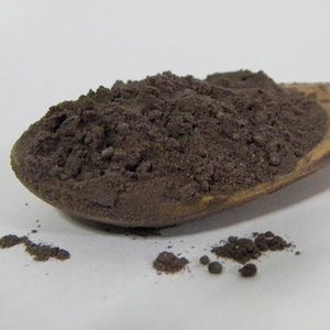 Op de afbeelding: Een houten lepel gevuld met een donkerbruin poeder. Het poeder is verspreid over een wit oppervlak.