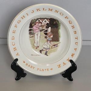 May include: A white ceramic baby plate with a gold rim and the alphabet printed around the edge. The centre of the plate features a colourful illustration of two children playing in a yard. The text "Baby Plate" is printed on the bottom of the plate.