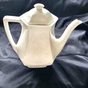 Può includere: Teiera in ceramica bianca con coperchio e corpo esagonale. La teiera ha un beccuccio e un manico.