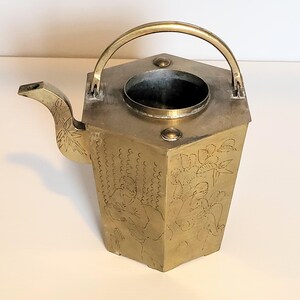 Puede incluir: Una tetera de latón con un diseño grabado de personas y flores. La tetera tiene un cuerpo hexagonal y un pico curvo.