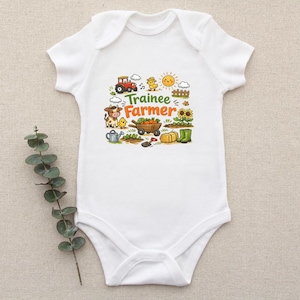 May include: A white short-sleeved baby bodysuit featuring a colorful graphic print with the text "Trainee Farmer". The illustration includes a red tractor, a cow, a chick, a wheelbarrow with vegetables, a sun, sunflowers, and farm tools.