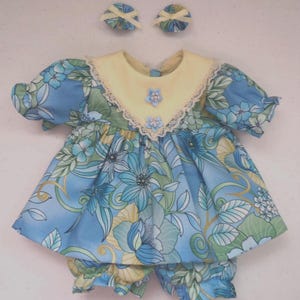May include: A doll dress featuring a blue and green floral pattern with a pale yellow collar, lace trim, and two small blue flower embellishments. The set includes matching bloomers and two small circular hair accessories with cream-colored ribbons.