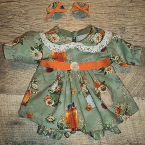 May include: A doll dress and two matching hair bows. The dress is light green with a floral pattern, a white lace collar, and an orange belt. The bows are green with orange ribbons. The outfit is designed for a doll.