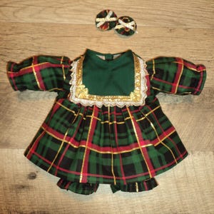 May include: A doll outfit featuring a green, red, and black plaid dress with gold accents and matching bloomers. The dress has a green collar with gold trim and matching hair bows.