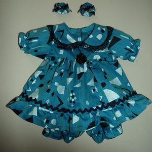 May include: A teal doll dress with a geometric pattern in black and white, featuring a black lace collar and a black flower accent. Matching teal and black patterned doll bonnets complete the set.