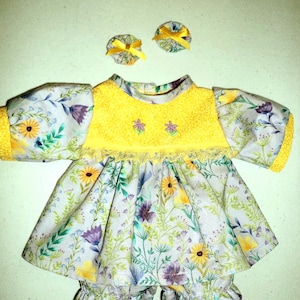 May include: A doll outfit consisting of a short-sleeved dress and matching bloomers featuring a floral print with yellow, purple, and green wildflowers on a white background. The dress has a yellow bodice with two small embroidered flowers.