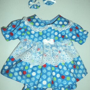 May include: A blue dress with a pattern of red, white, and green polka dots, featuring a white lace collar, a white lace ruffled waist overlay with a small white bow, and short puffed sleeves. Includes matching blue polka dot hair accessories.