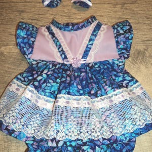 May include: A doll dress featuring a dark blue fabric with a light blue and purple leaf pattern, a solid light purple bodice panel, white lace trim, and a small ribbon rosette. The set includes matching bloomers and two small circular hair accessories.