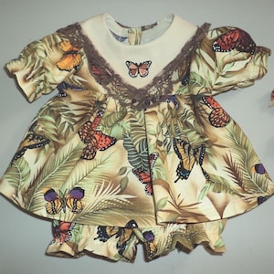 May include: A two-piece baby outfit featuring a butterfly and leaf print in shades of tan, green, orange, and purple. The set includes a short-sleeved dress with a cream-colored collar and matching ruffled shorts. Two matching hair bows are also included.