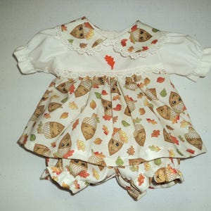 May include: A white baby outfit with a collared top and matching shorts. The outfit features a print of smiling acorns and autumn leaves in shades of brown, orange, and green. The collar and sleeves are trimmed with lace.