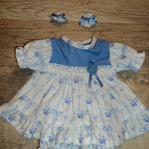 May include: A doll dress with a blue bodice and a white skirt featuring a blue floral and ribbon pattern. The dress has short puff sleeves, a white lace trim at the waist, a blue ribbon flower accent, and a ruffled hem, accompanied by two matching hair accessories.