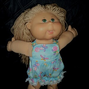 May include: A vintage doll with blonde yarn hair and green eyes. The doll wears a light blue dress with a butterfly pattern and white lace trim. The doll's arms are outstretched, and it has a smiling expression.