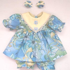 May include: A doll outfit set featuring a blue floral print dress with a yellow collar and matching bloomers. The dress has a lace trim and is adorned with blue flower accents. Two matching hair bows complete the set.