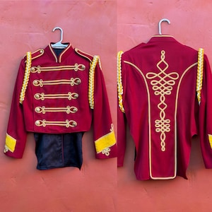 Puede incluir: Chaqueta de banda de música burdeos con ribetes y botones dorados. La chaqueta tiene detalles amarillos en las mangas y los hombros. La parte trasera de la chaqueta presenta un elaborado diseño bordado en oro.