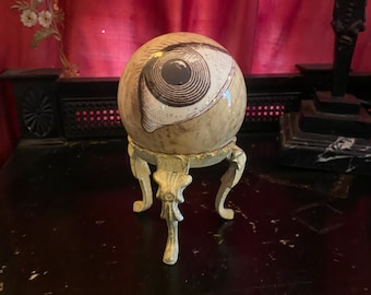 May include: A spherical object featuring a detailed black and white illustration of a human eye on its surface. The sphere is supported by a decorative, light-colored metal stand with three ornate legs, providing a stable base for the round item.
