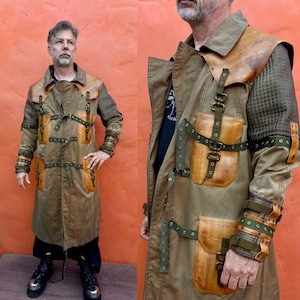 May include: A long olive green coat featuring tan leather panels on the chest and shoulders, multiple olive green straps with metal buckles, and textured dark green sleeves. The coat includes large tan leather pockets and metal eyelet details.