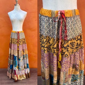 May include: A patchwork maxi skirt in shades of yellow, purple, and brown, displayed on a mannequin. The skirt features a drawstring waist and a tiered design with various patterned fabric panels. The skirt has a bohemian style.