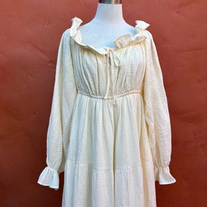 May include: A cream-colored long-sleeved dress featuring a textured fabric, a ruffled neckline with a central tie closure, an elasticized waist, and tiered skirt detailing. The garment has long sleeves that end in ruffled cuffs, providing a full silhouette.