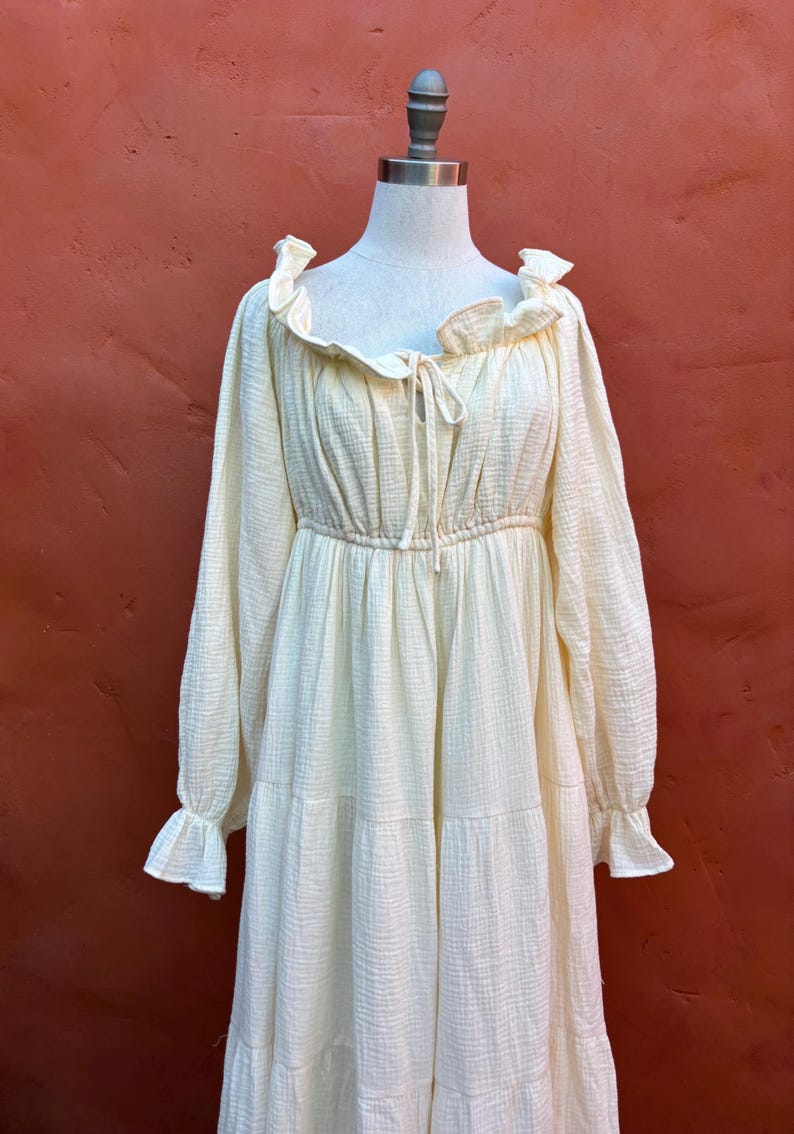 May include: A cream-colored long-sleeved dress featuring a textured fabric, a ruffled neckline with a central tie closure, an elasticized waist, and tiered skirt detailing. The garment has long sleeves that end in ruffled cuffs, providing a full silhouette.