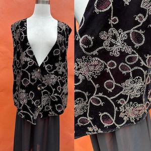 May include: A black velvet vest featuring a repeating pattern of gold-toned chains and stylized floral motifs with deep red accents. The sleeveless garment has a deep V-neckline and is secured at the front with three round, metallic-colored buttons.
