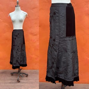 May include: A long, black skirt with a unique design, displayed on a mannequin. The skirt features a textured fabric with floral embroidery and a contrasting black velvet panel. A ruffled hem adds a decorative touch.