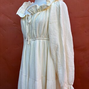 May include: A pale yellow long-sleeved dress made of a textured muslin fabric. The dress features a ruffled neckline with a drawstring tie, a gathered waistline, and long sleeves that end in ruffled cuffs. The skirt has a tiered design.
