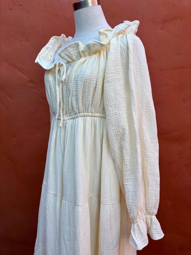 May include: A pale yellow long-sleeved dress made of a textured muslin fabric. The dress features a ruffled neckline with a drawstring tie, a gathered waistline, and long sleeves that end in ruffled cuffs. The skirt has a tiered design.