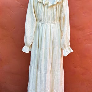 May include: A long-sleeved, cream-colored maxi dress featuring a textured, crinkled fabric. The garment includes a ruffled neckline, an elasticized waist, and tiered skirt sections. The sleeves are finished with ruffled cuffs, creating a gathered look.