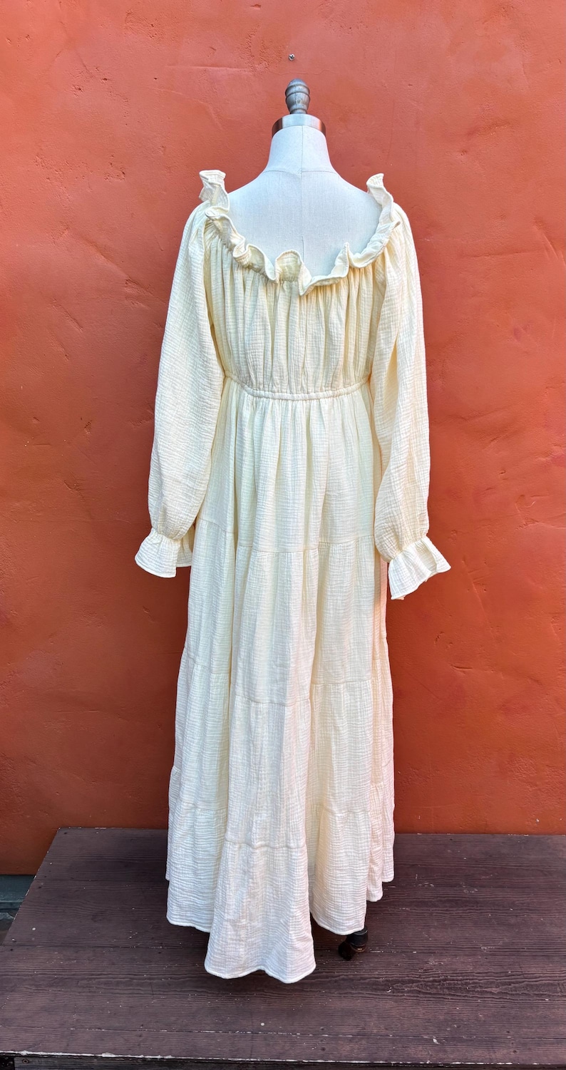 May include: A long-sleeved, cream-colored maxi dress featuring a textured, crinkled fabric. The garment includes a ruffled neckline, an elasticized waist, and tiered skirt sections. The sleeves are finished with ruffled cuffs, creating a gathered look.